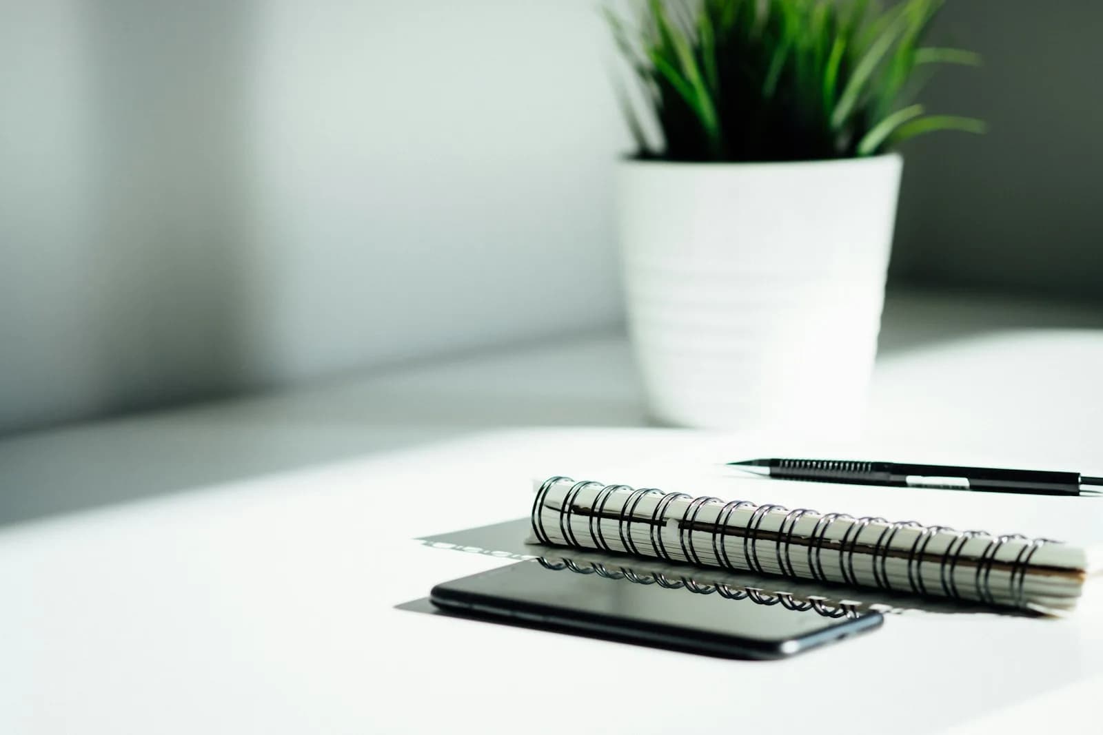 A notebook and pen beside a phone on a sunlit desk.